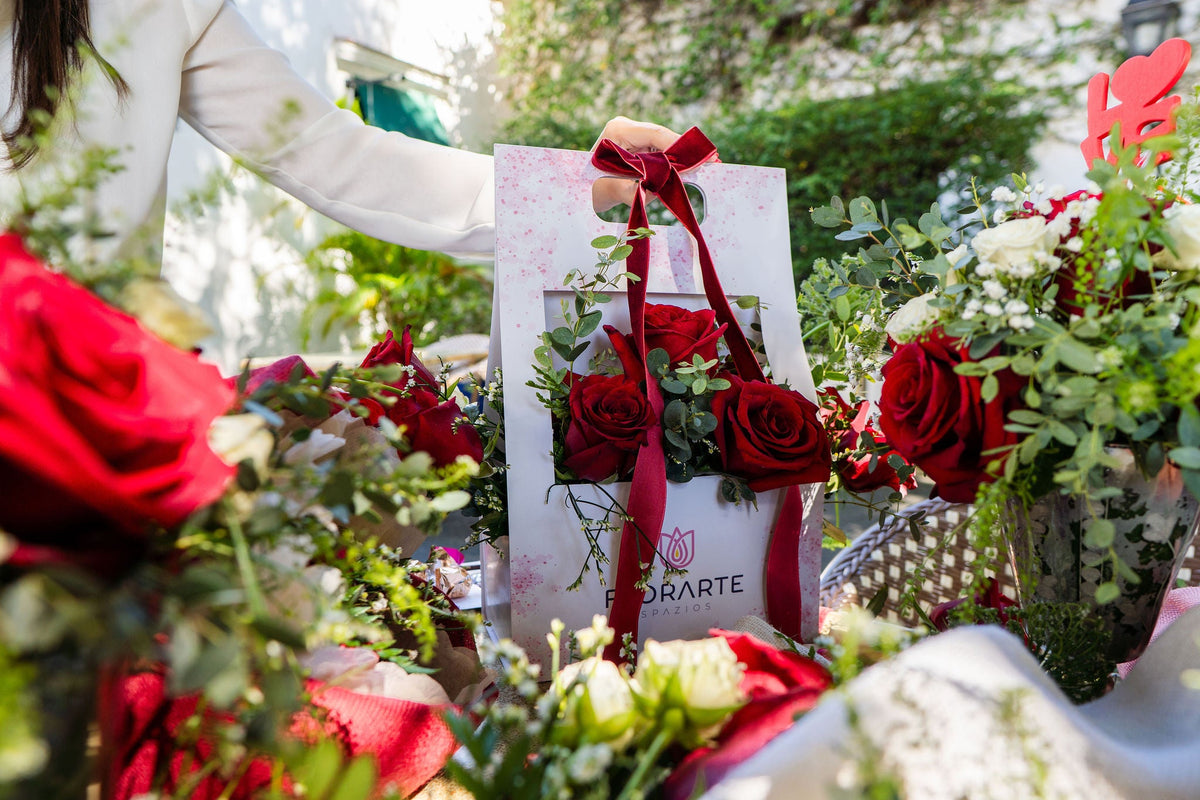 Una mano entrega una bolsa blanca de Florarte con un lazo rojo, de la que emergen rosas rojas y follaje verde. Alrededor, arreglos florales sobre una mesa al aire libre, iluminados por la luz del día, crean una escena romántica y elegante.